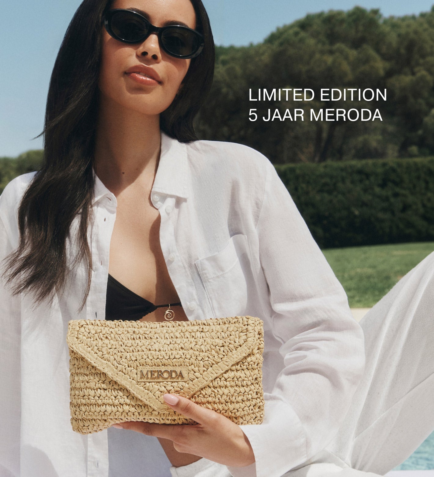 Summer Travel Clutch van geweven papierstro vastgehouden door een vrouw in een wit linnen shirt en zonnebril.